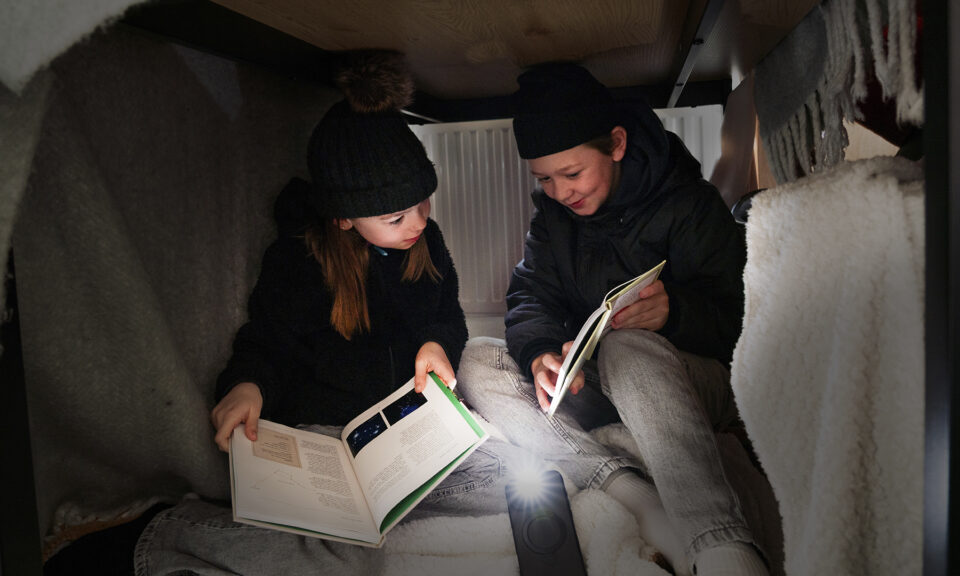 Två barn med mössa och varma kläder sitter i en koja av filtar och läser böcker i ljuset av mobilens lampa.